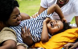 a two parents and their child smiling and playing together on a blanket on the grass outside
